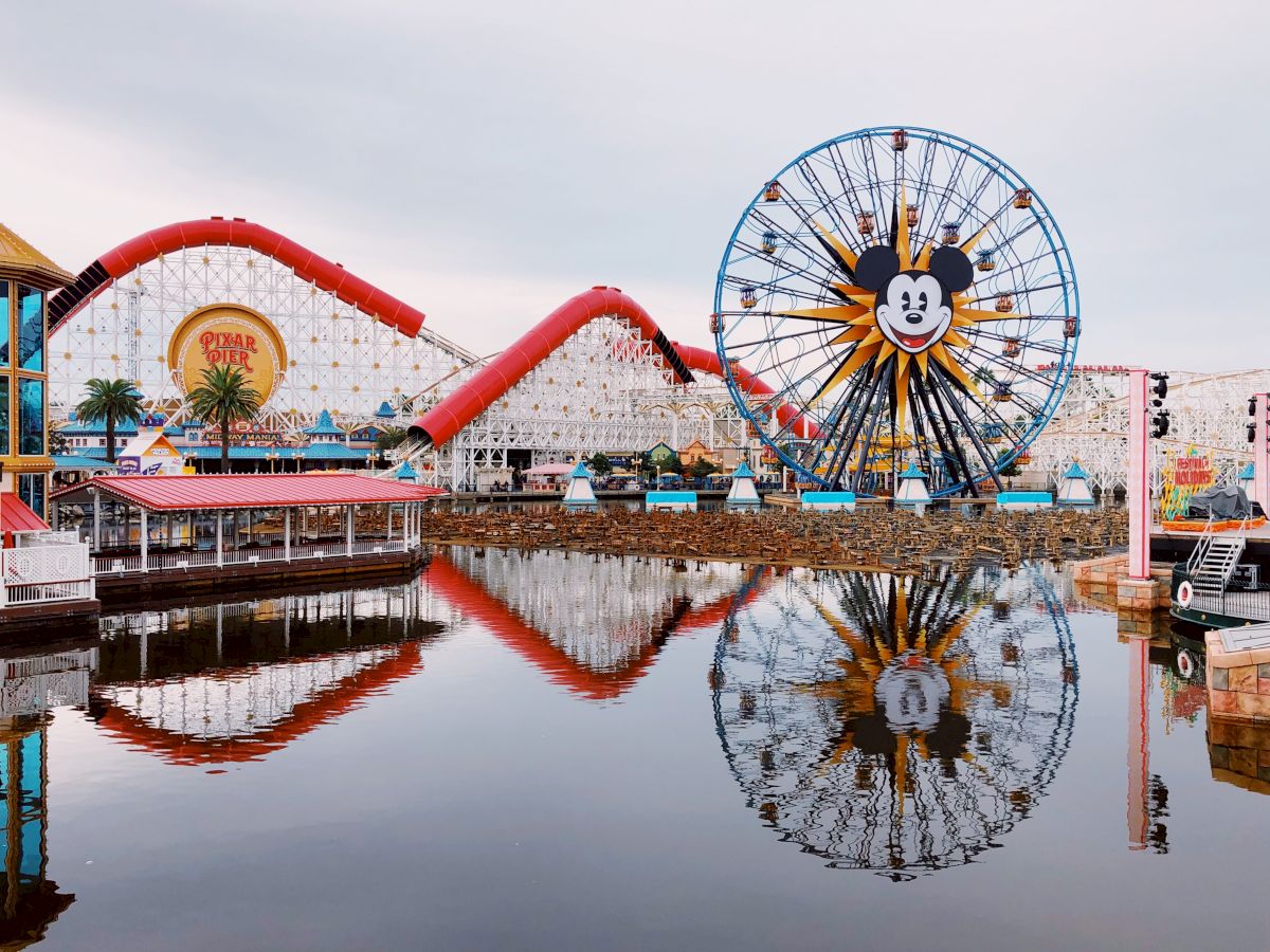 A theme park with a large Ferris wheel featuring Mickey Mouse, a roller coaster, and other attractions is reflected in a calm body of water.