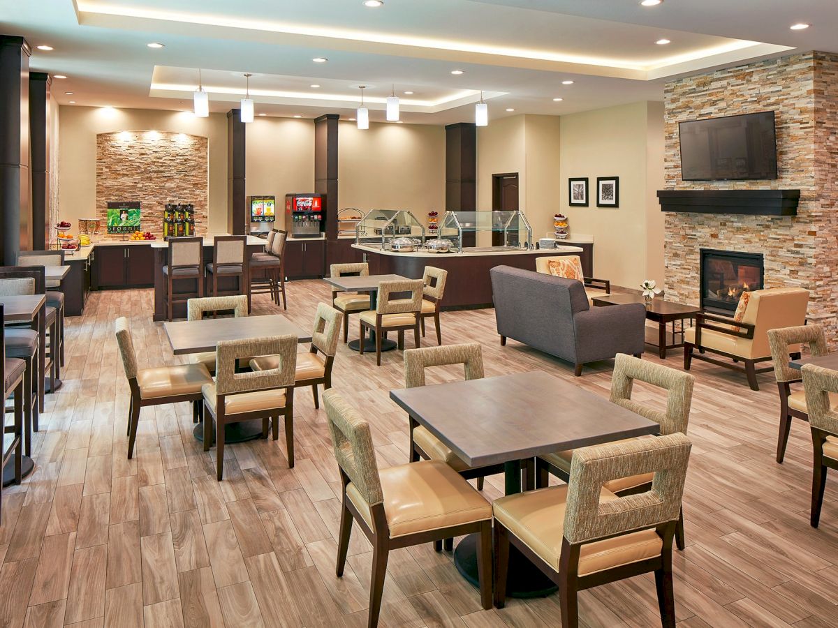 A modern dining area with wooden flooring, tables, chairs, a fireplace, and a self-serve food and beverage station, well-lit with ample seating.