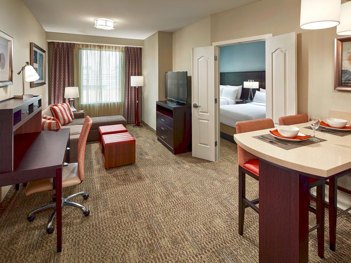 The image shows a modern hotel suite with a living area, a workspace, a dining area, and a separate bedroom with a bed visible through open doors.