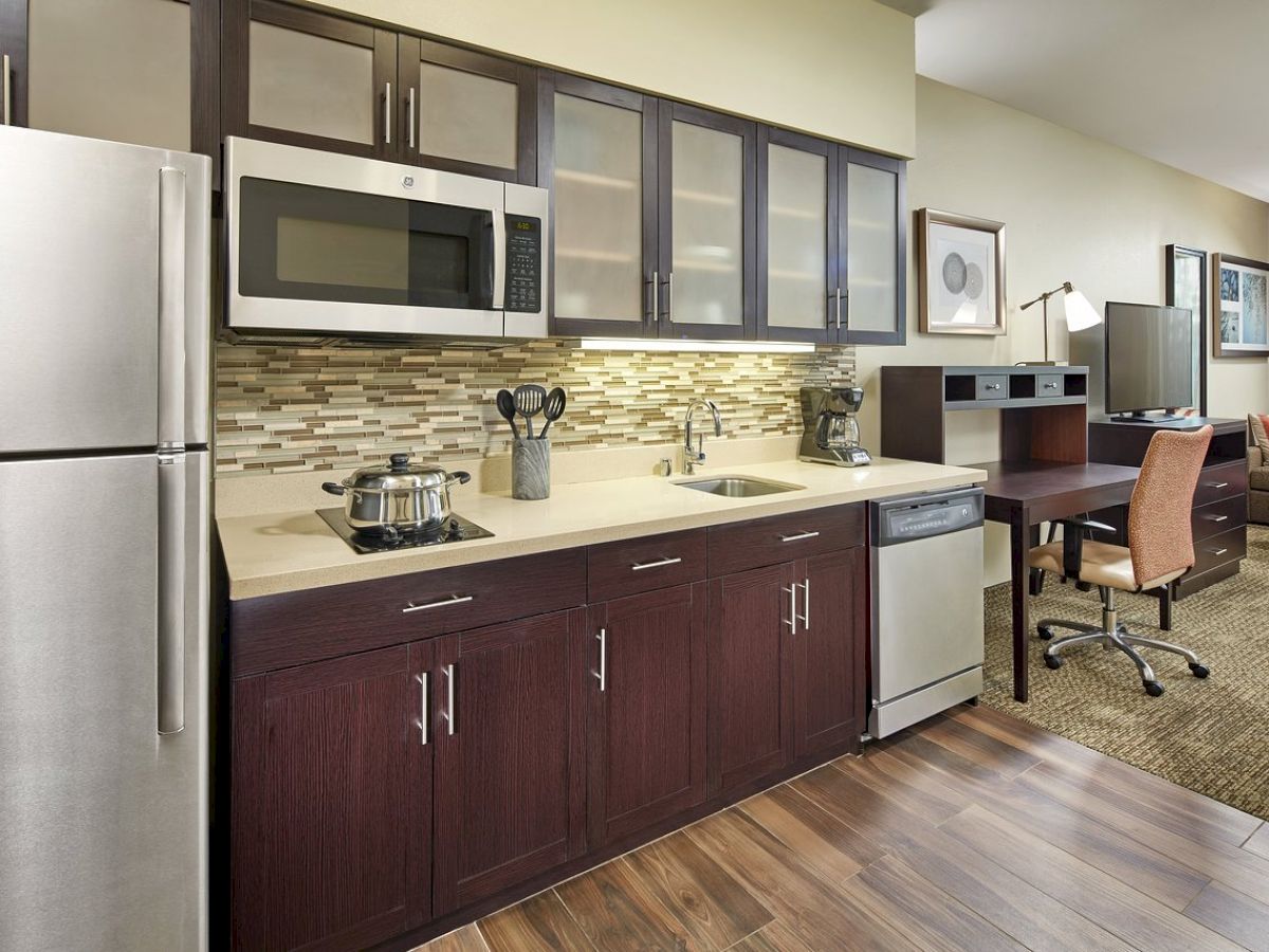 The image shows a modern kitchen area with stainless steel appliances, dark wood cabinets, a small desk setup, and a seating area in the background.