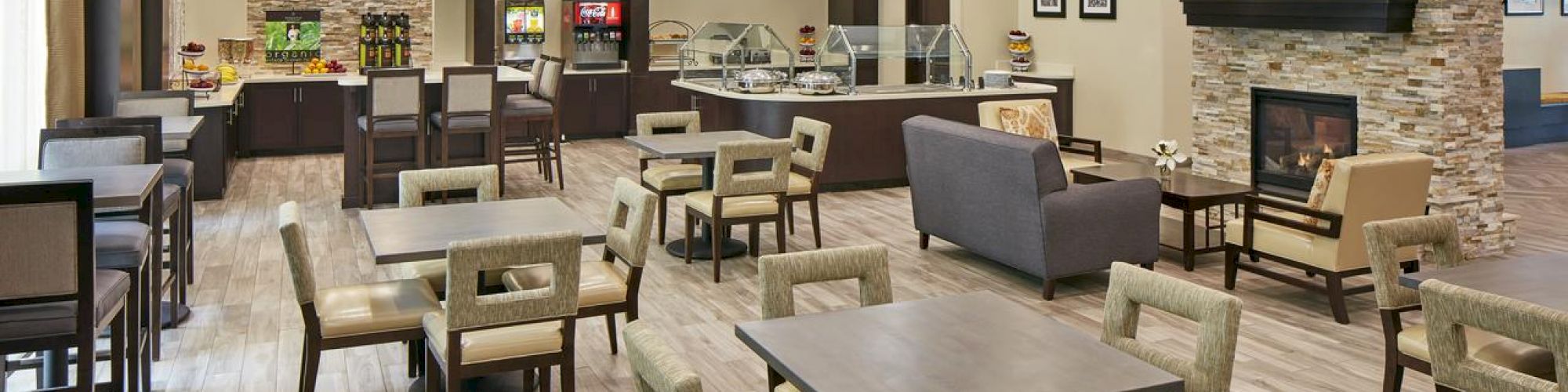 This image shows a modern dining or lounge area with tables, chairs, a fireplace, a TV, and a food service counter in the background.