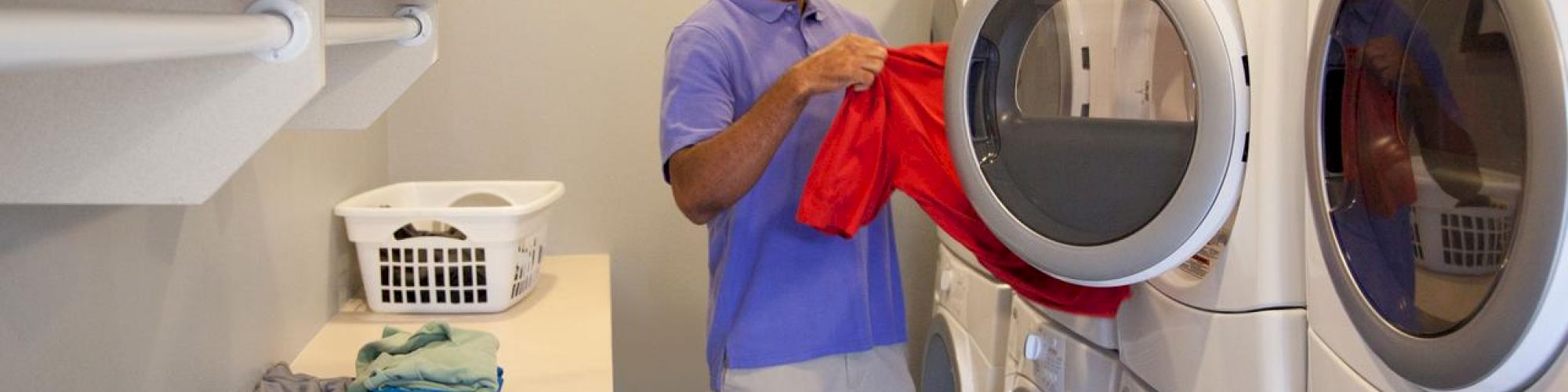 A man wearing a purple shirt is doing laundry, placing a red garment into a stacked washer-dryer in a laundry room with shelves.