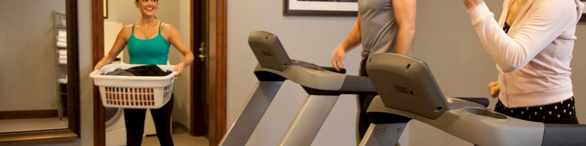 Three people in a home gym. Two are on treadmills smiling; one brings laundry. There’s a mirror, clock, and framed picture on the wall.