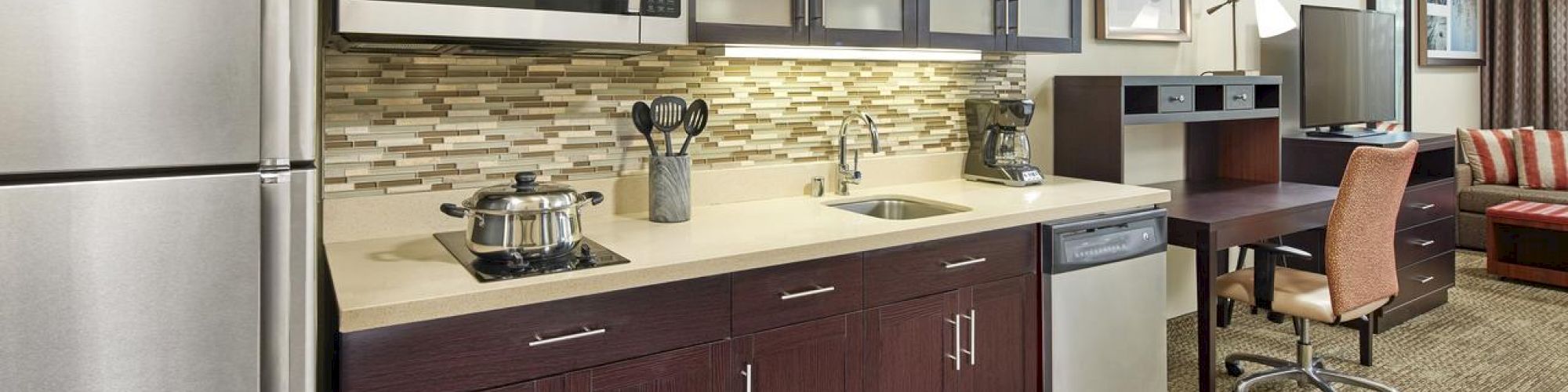 Modern kitchen with stainless steel appliances, dark wood cabinets, a microwave, sink, dishwasher, and a desk with a chair in the background.