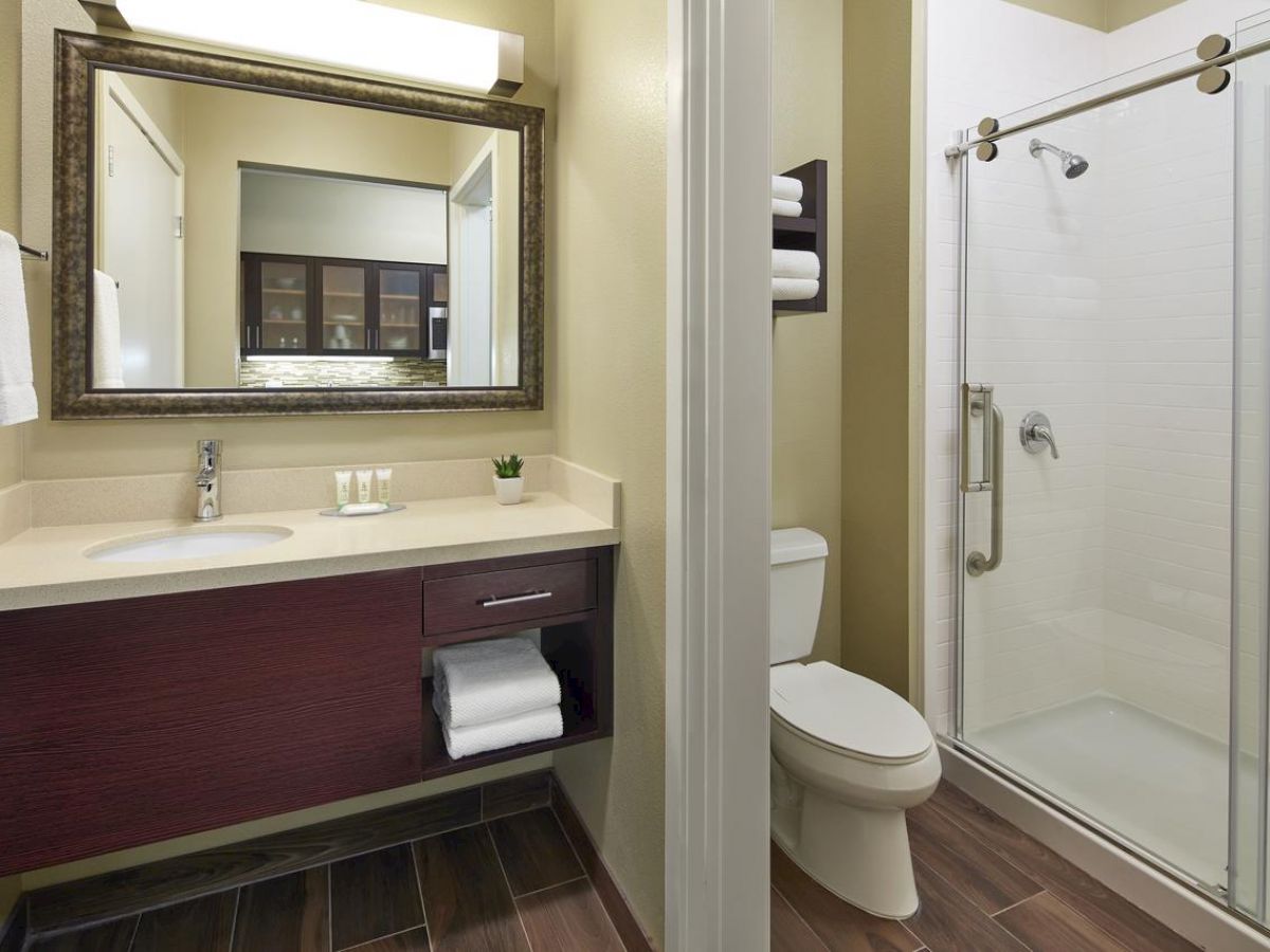 The image shows a modern bathroom with a sink and mirror, a toilet, and a glass-enclosed shower with towels on the shelf above the toilet.