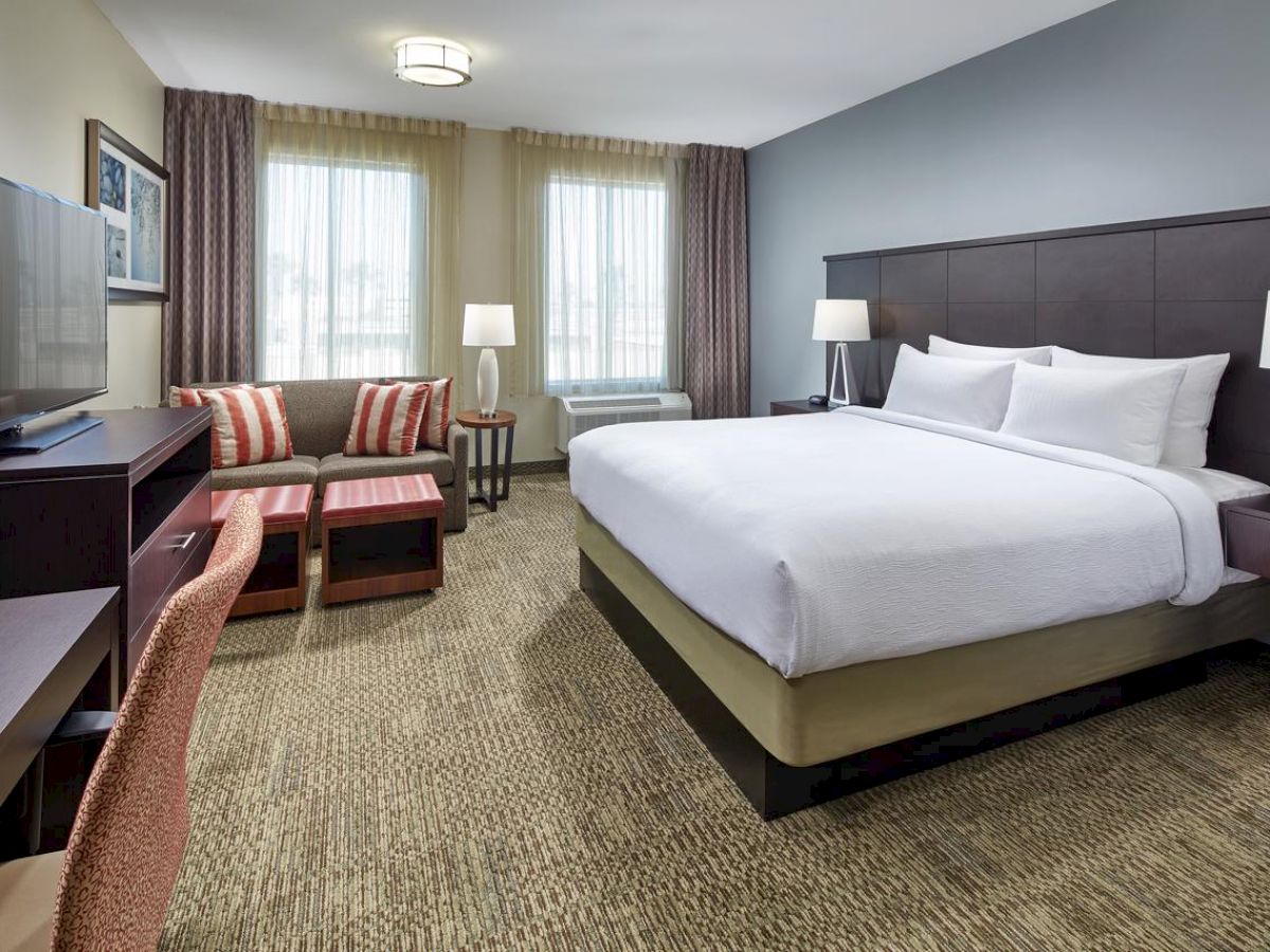 This image shows a modern hotel room with a large bed, desk, chair, TV, sofa, two lamps, and two windows with curtains, featuring neutral tones.