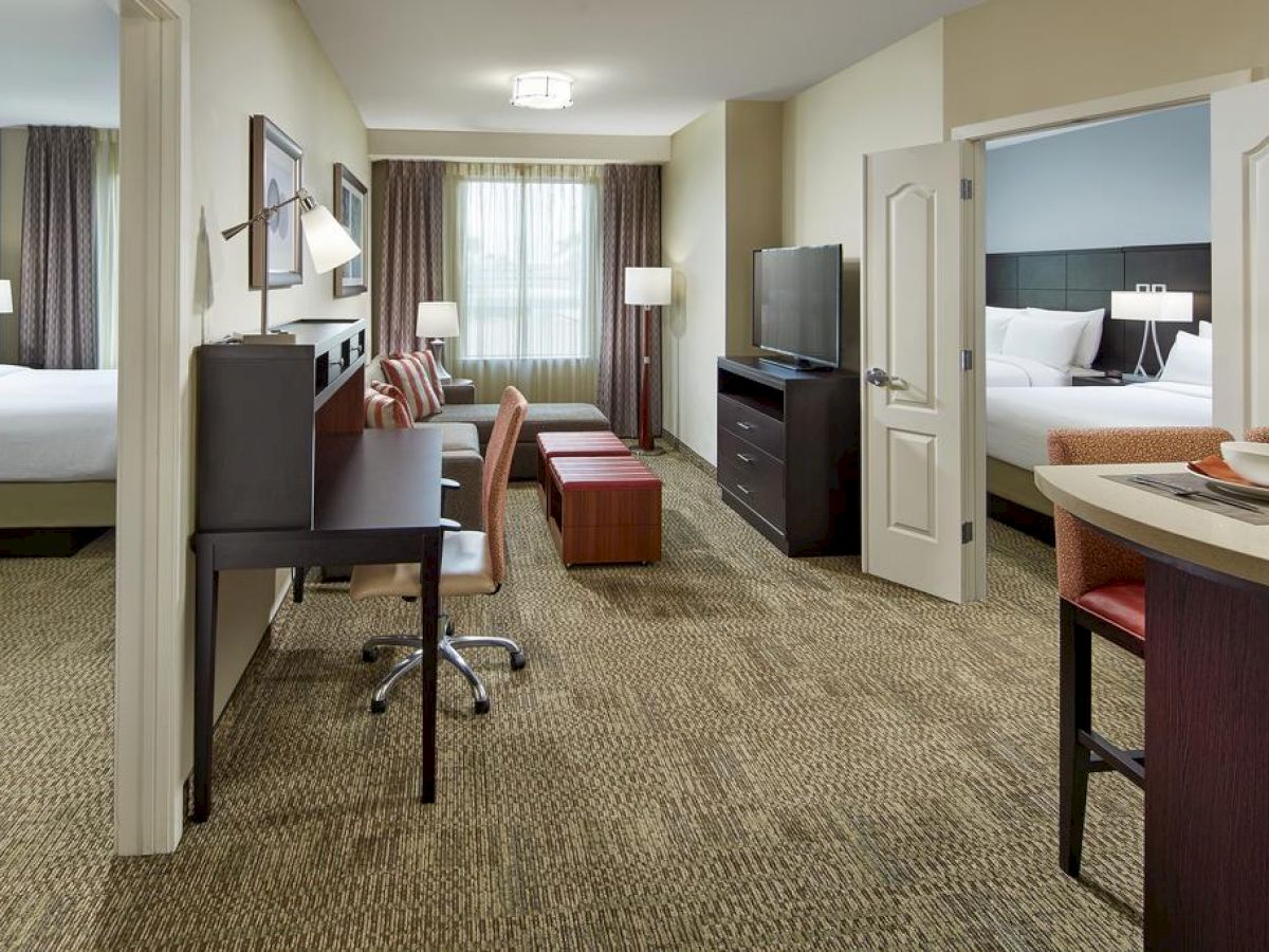 The image shows a hotel suite featuring two beds, a TV, desk, and seating area, separated by a partial wall. The room is well-lit and modern.