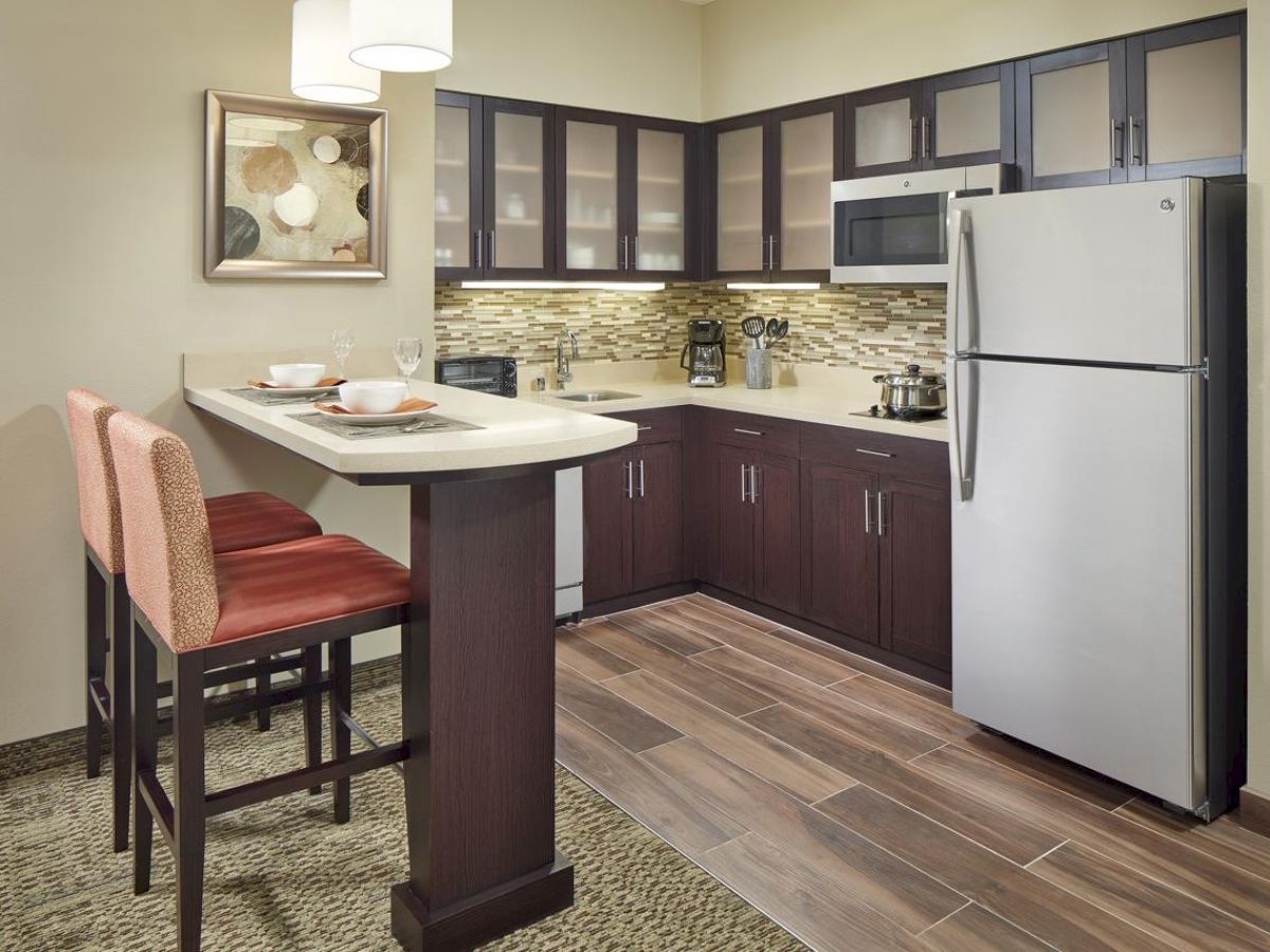 A modern kitchen with dark cabinets, a silver refrigerator, microwave, and a breakfast bar with two chairs. Wall art and pendant lights complete the look.