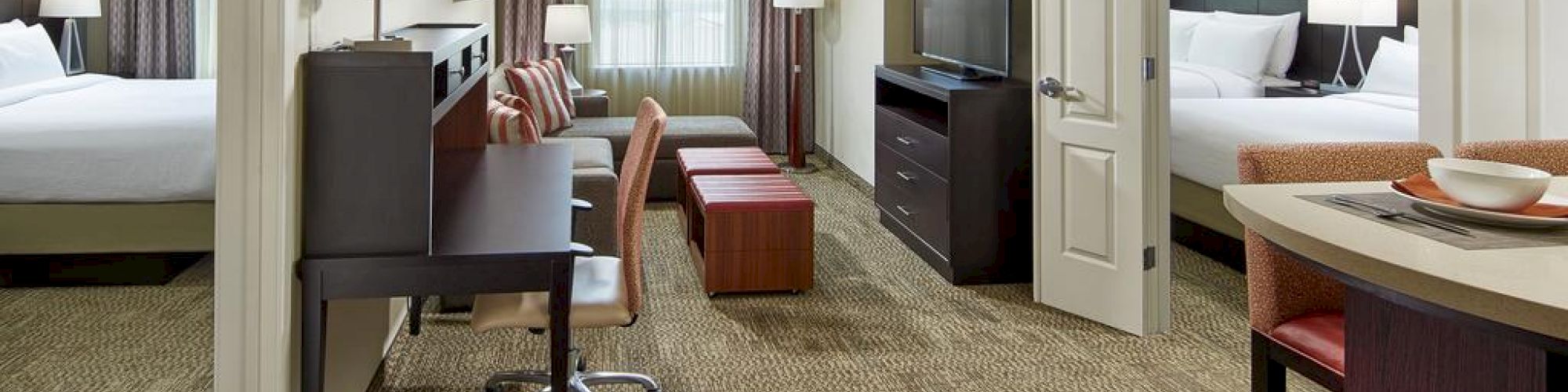 This image shows a hotel suite with two beds, a TV stand, a desk chair, curtains, and a small dining area with red chairs and a countertop.