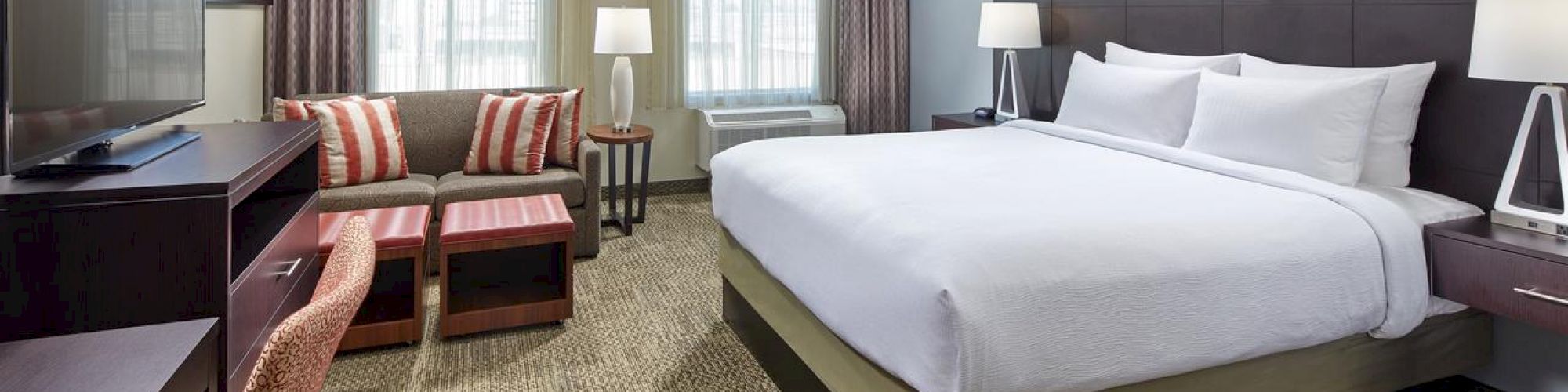 A neatly arranged hotel room with a king-sized bed, TV, desk, chair, sofa, and coffee table. The room features large windows with curtains and lamps.