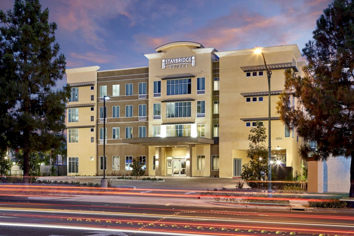 This image shows an exterior view of a Staybridge Suites hotel building at dusk, with light trails from passing cars in the foreground ending the sentence.