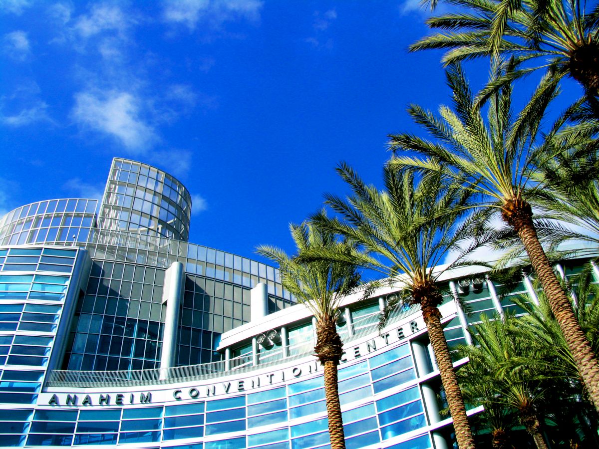 The image shows a modern glass building with "Anaheim Convention Center" on its facade, set against a blue sky with palm trees in the foreground.