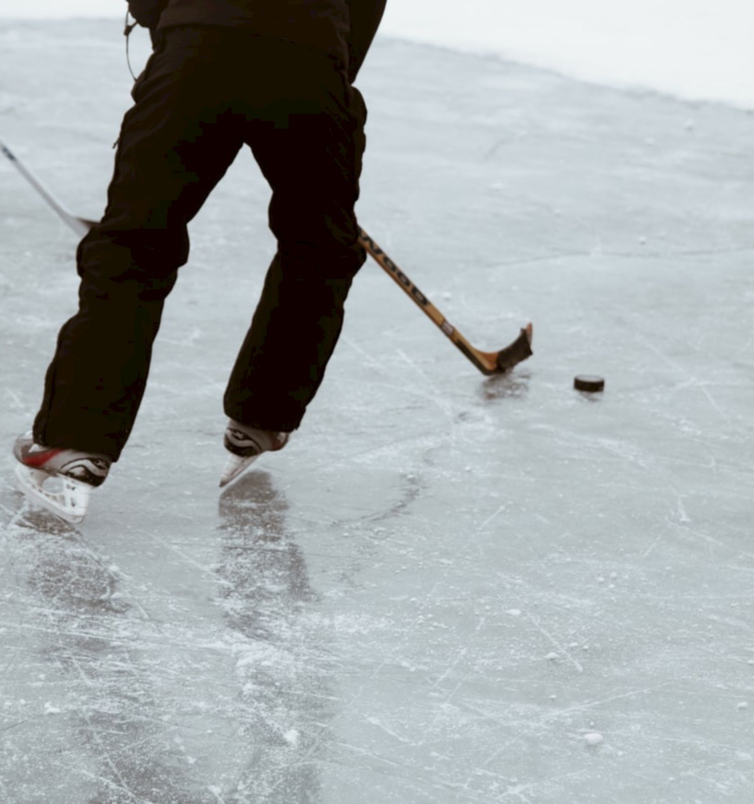 Two people are playing hockey on an ice rink, focusing on the puck and using their hockey sticks to maneuver.