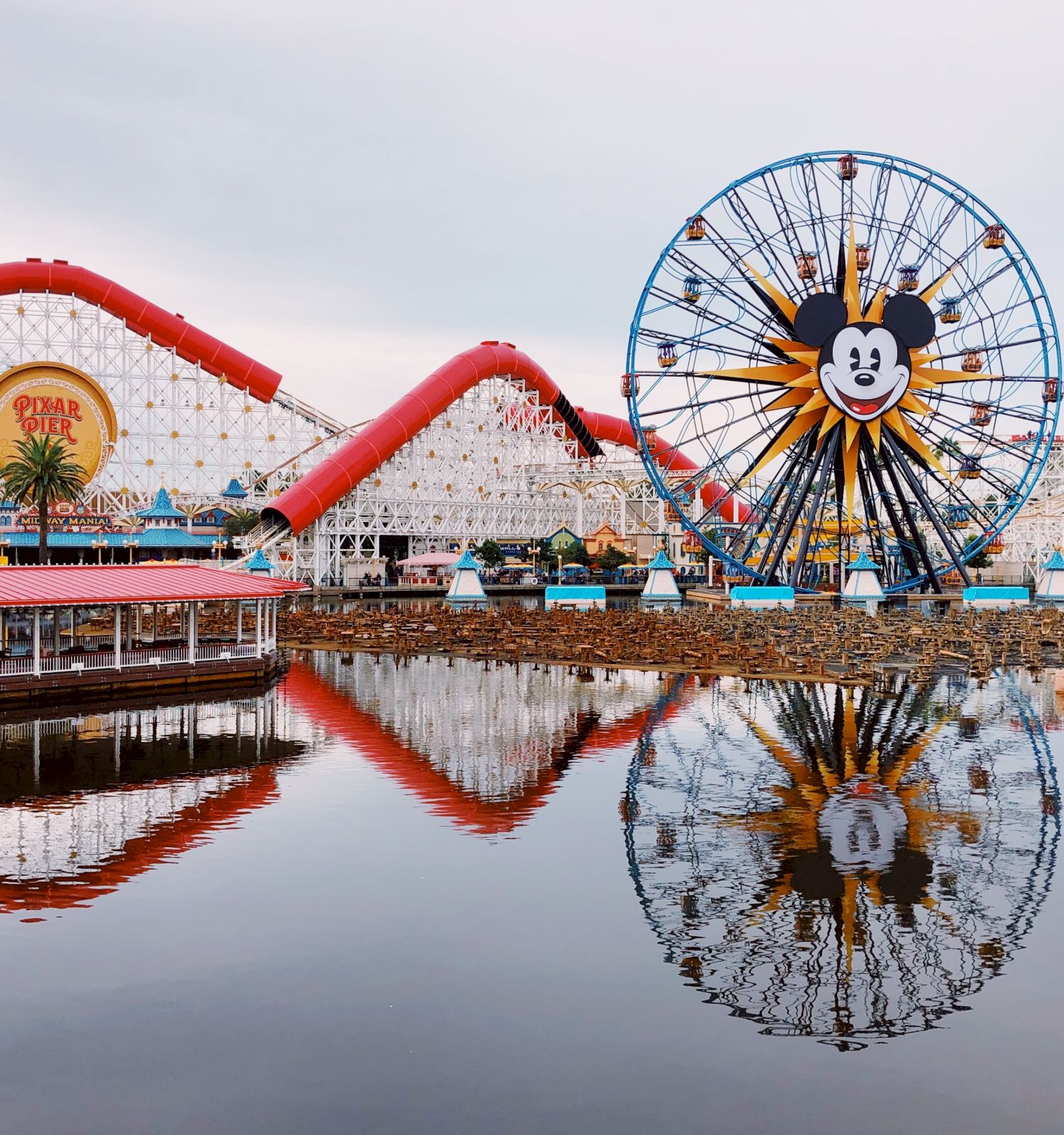 The image shows a theme park featuring a Ferris wheel and roller coaster with reflections in a body of water, creating a lively and colorful atmosphere.