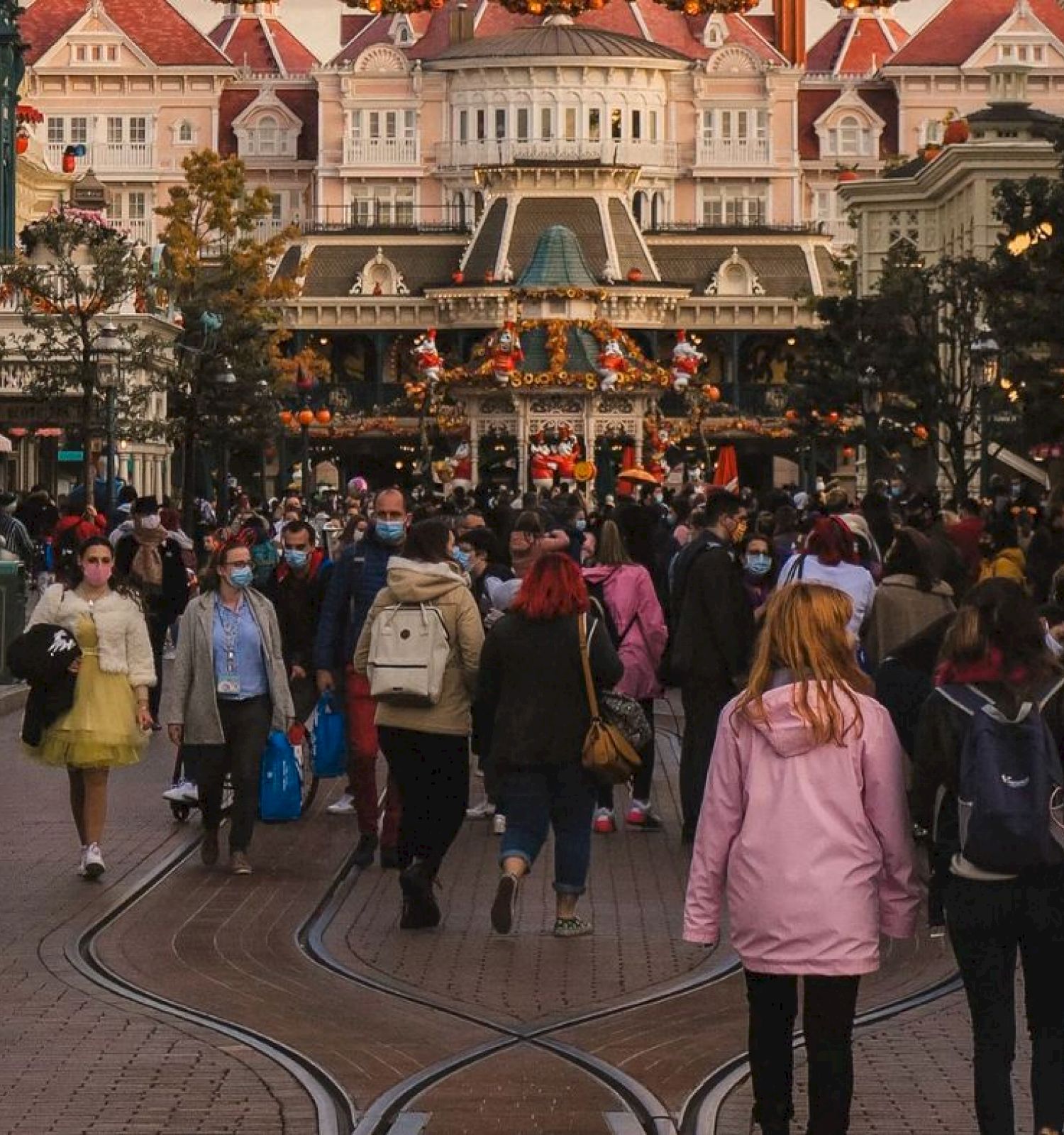 A large crowd of people is walking through a lively, decorated street with a grand building in the background at what appears to be a theme park.