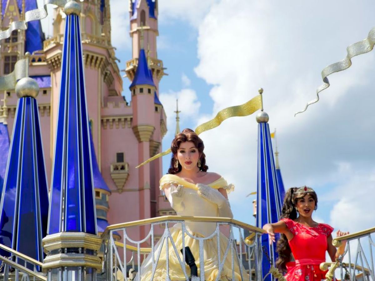 Two costumed characters pose in front of a castle with pointed blue and gold accents, under a partly cloudy sky.