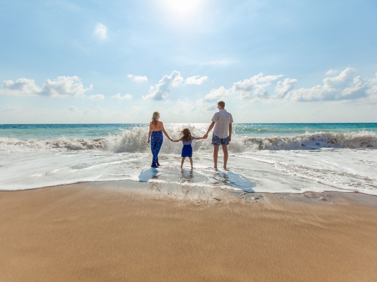 A family of three is holding hands and standing at the edge of the ocean, facing the sea, on a sunny day with a clear sky.