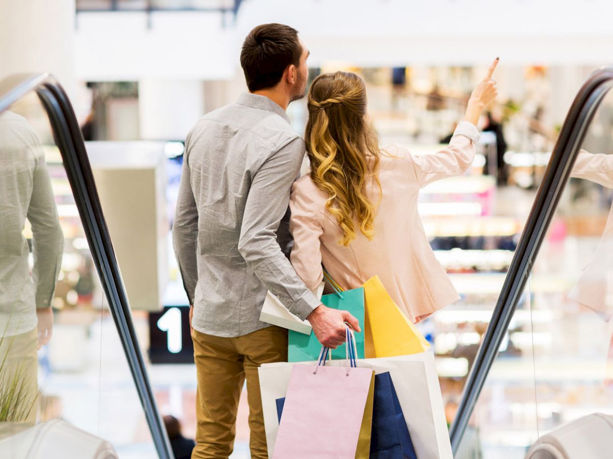 A couple holding shopping bags stands on an escalator in a mall; the woman is pointing ahead while they are in mid-conversation.