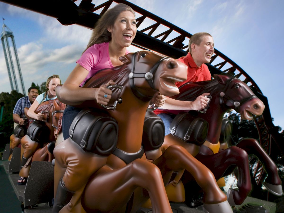 People are riding a roller coaster with horse-shaped seats, enjoying the thrill of the ride under a blue sky with some clouds in the background.