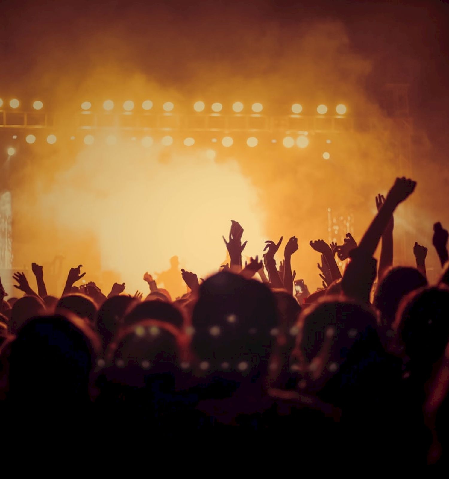 A crowd is at a concert with vibrant stage lights in the background, raising their hands and enjoying the live music performance.