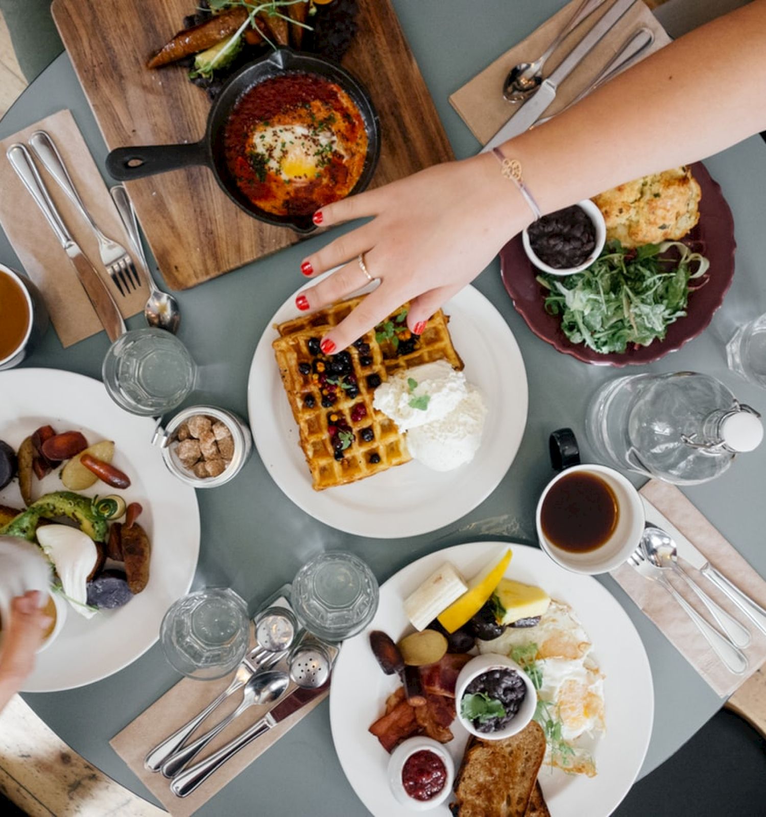 A table with various delicious dishes including waffles, bread, and salads, surrounded by drinks and hands reaching for food and beverages.