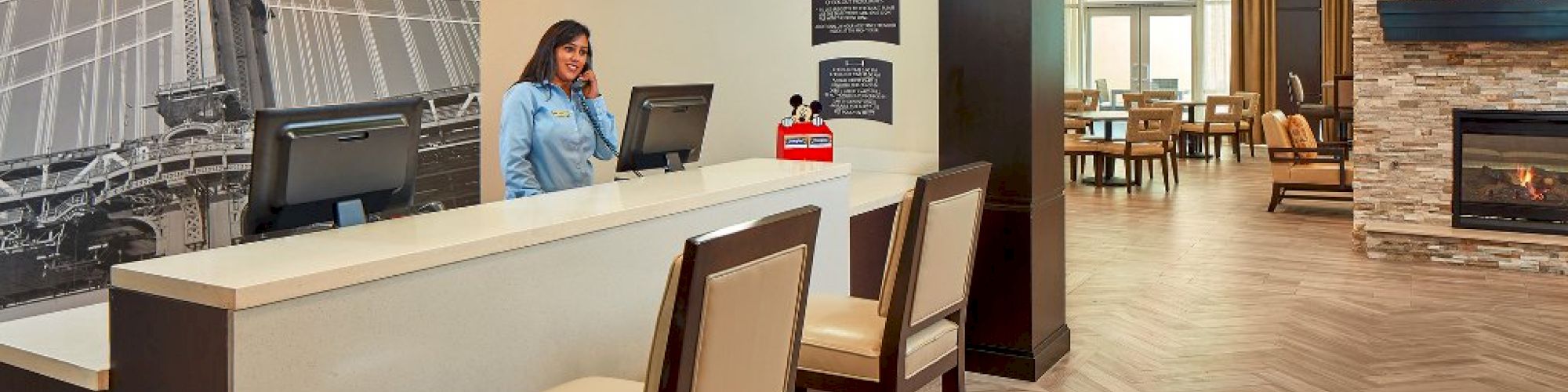 A woman stands at a reception desk in a modern, warm-toned lobby with chairs and a fireplace in the background.