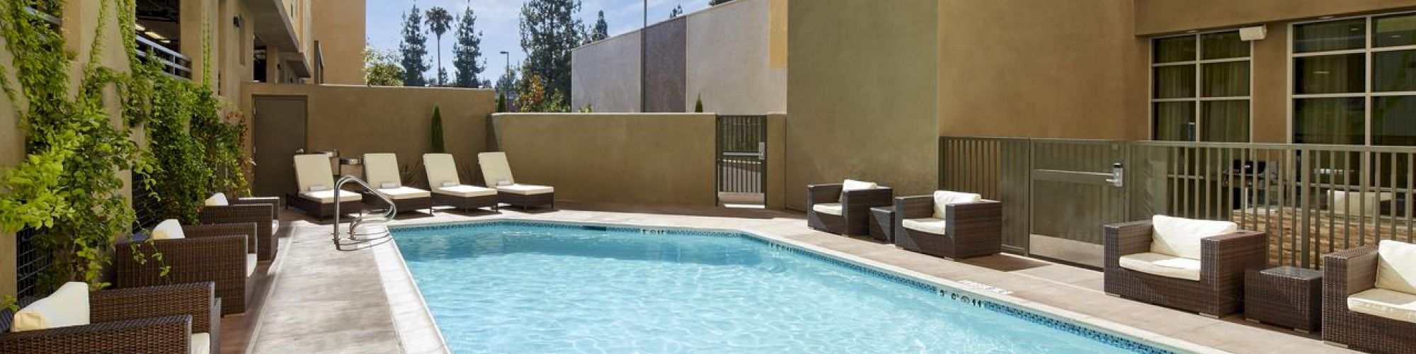 The image features a small outdoor pool surrounded by lounge chairs and cushioned outdoor furniture, located between tall buildings under a clear sky.