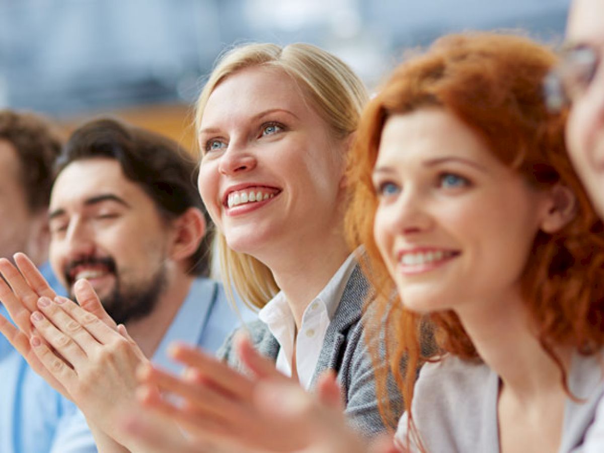 A group of people sitting in a row, smiling and clapping, possibly during a meeting or seminar, shown in an indoor setting.