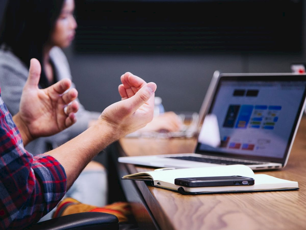 A person in a plaid shirt is gesturing near an open laptop on a desk, with another person blurred in the background.