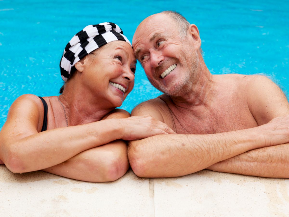 An older couple, smiling, rests at the edge of a swimming pool, with the woman wearing a striped swim cap.