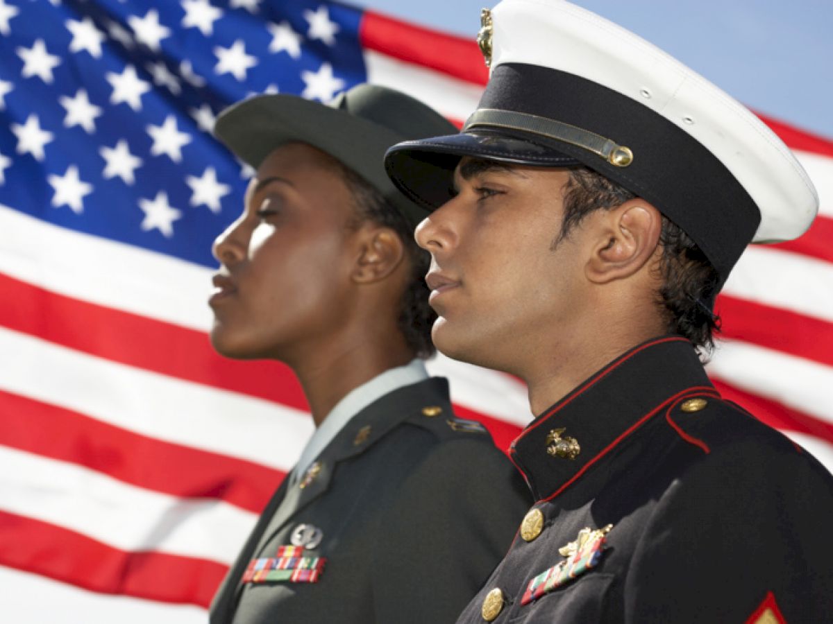 Two military officers in uniform stand in front of an American flag, looking forward.