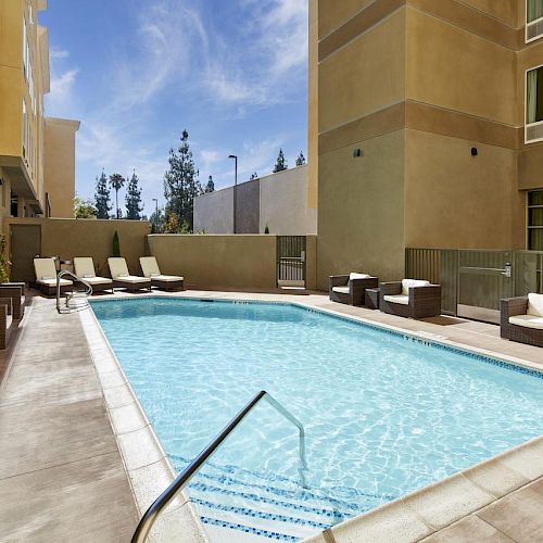 The image shows an outdoor swimming pool surrounded by lounge chairs and cushioned seating areas, set between modern buildings under a clear blue sky.