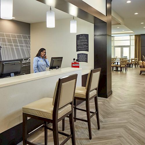 A reception area with a woman at the desk, two chairs in front, modern decor, wall art, and a seating area with a fireplace in the background.