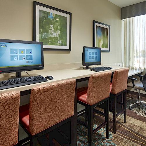 The image shows a small office area with two desktop computers on a long desk, three chairs, and a window with blinds and curtains.