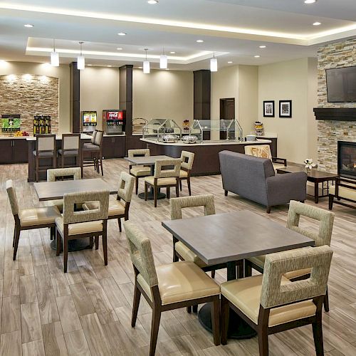 The image shows a modern dining area with wooden flooring, multiple tables and chairs, a fireplace, a TV, and a countertop with food and drink options.