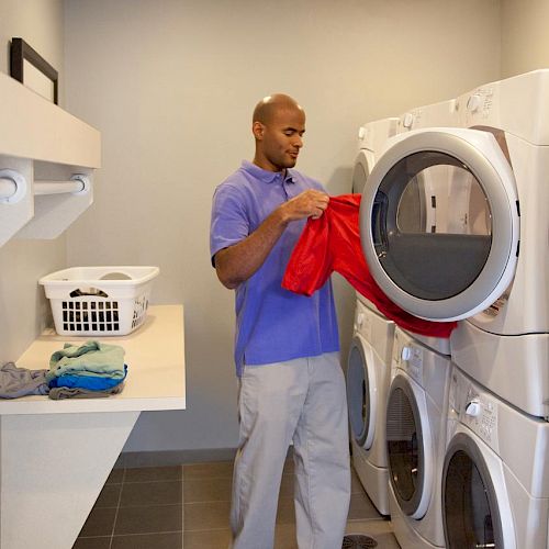 A person is doing laundry in a room with stacked white washers and dryers, folding red clothes next to a countertop holding a laundry basket and folded items.