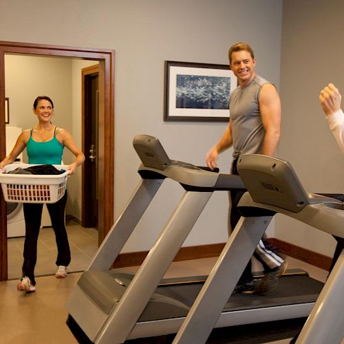 Three people are in a gym: two using treadmills while a third, holding a laundry basket, walks in.