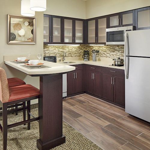 A modern kitchen with dark wood cabinets, a stainless steel fridge, a microwave, and a breakfast bar with two red chairs.