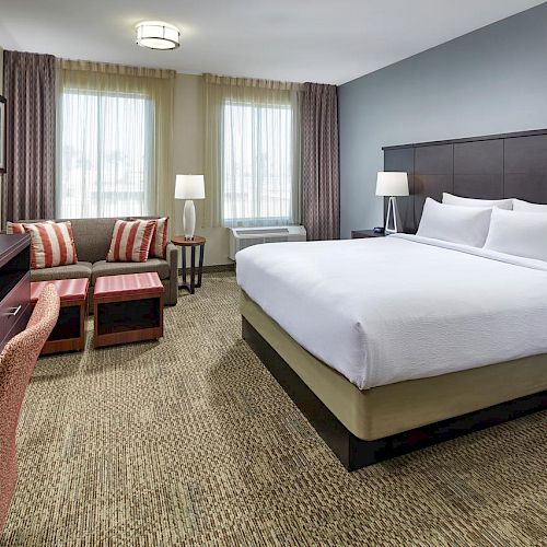 A cozy hotel room featuring a large bed, a sofa, a television, a desk, nightstands, and lamps. The room has carpeted floors and window curtains.