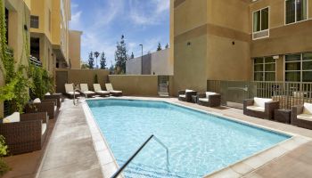 This image shows a modern outdoor swimming pool surrounded by lounge chairs and wicker seating in a courtyard of an apartment or hotel.