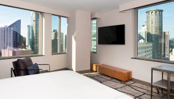 A modern room with large windows showing city skyline, a wall-mounted TV, armchair, small table, and bed in view, ending the sentence.