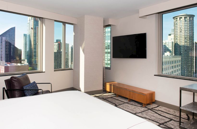A modern room with large windows showing city skyline, a wall-mounted TV, armchair, small table, and bed in view, ending the sentence.