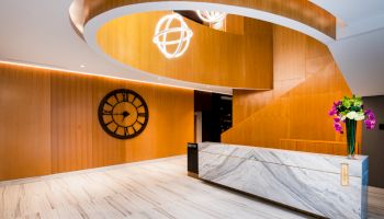 This image shows a modern reception area with a marble desk, a large wall clock, wooden accents, a spiral staircase, and a vase of flowers.