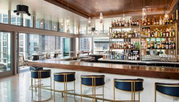 A modern bar with a wooden top, gold and navy blue barstools, and shelves full of various liquor bottles. Glass windows offer a city view.