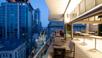 A modern high-rise balcony with outdoor seating overlooks a cityscape with tall buildings and a street lined with lights, captured at dusk.