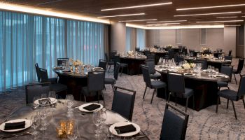 The image depicts an elegantly set dining hall with round tables, black chairs, and place settings including glassware and cutlery, ready for an event.