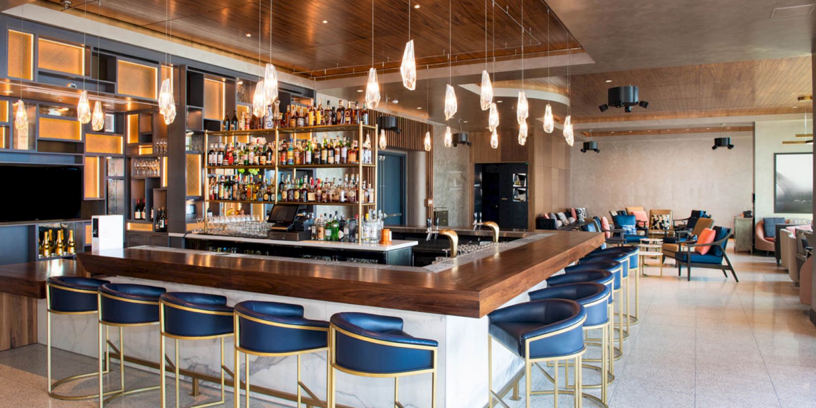 The image features a modern bar with a fully stocked back shelf, stylish barstools, hanging lights, and a relaxed seating area in the background.
