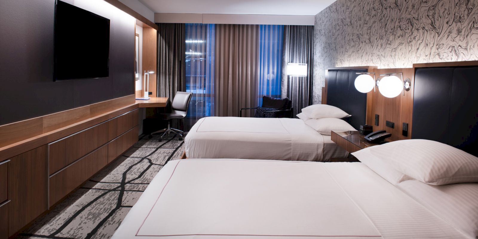 The image shows a modern hotel room with two neatly made beds, a TV mounted on the wall, a work desk, and stylish decor.