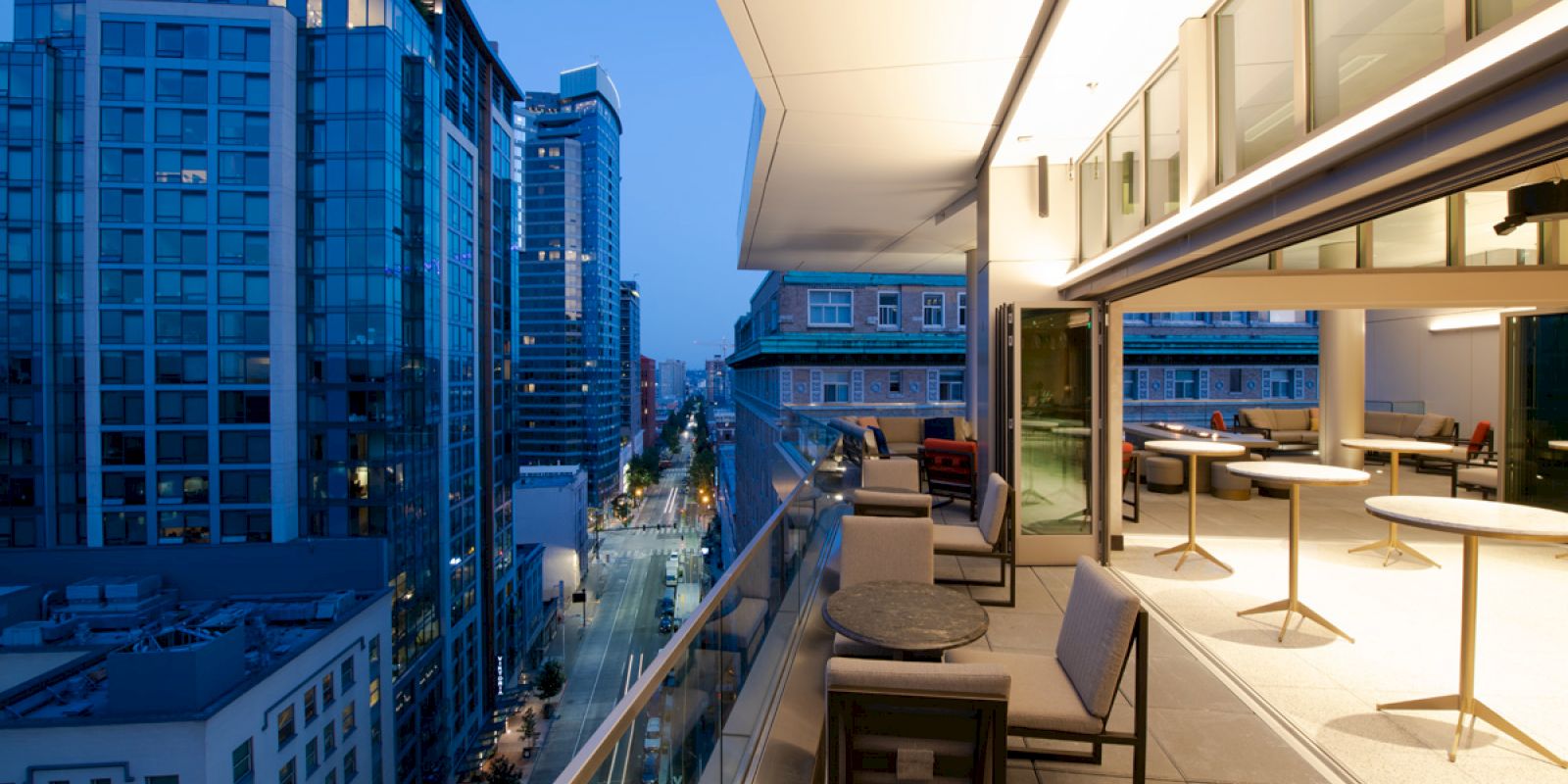 A modern cityscape with tall buildings, viewed from a stylish rooftop terrace with seating, tables, and glass railings during evening hours.