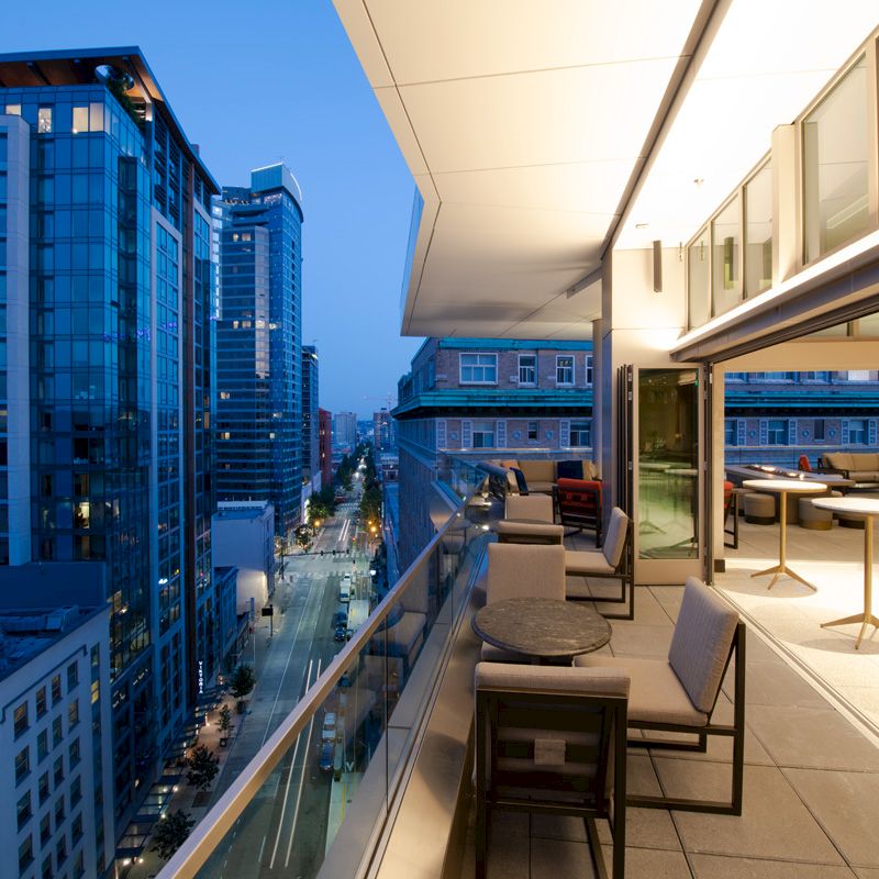 The image shows a modern balcony with seating, overlooking a city street lined with tall buildings. The interior looks stylish and well-lit.